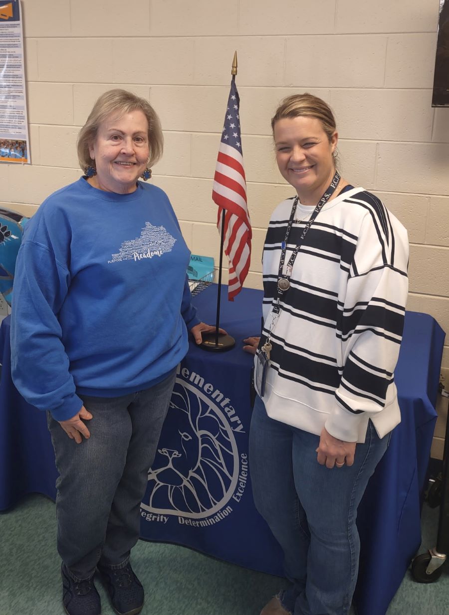 Two women stand on either side of a flag, which stands about 2 feet tall. The flag is on top of a table with a blue tablecloth.