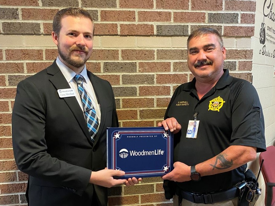 Two men are pictured. On the left is WoodmenLife Representative Casey Morehead; he is wearing a suit and a name tag. On the right is School Resource Officer Ronnie Harvell; he is wearing a dark polo shirt with his name and the police department logo. They are holding either side of a navy blue box that says "Proudly presented by WoodmenLife." Inside is an American flag.