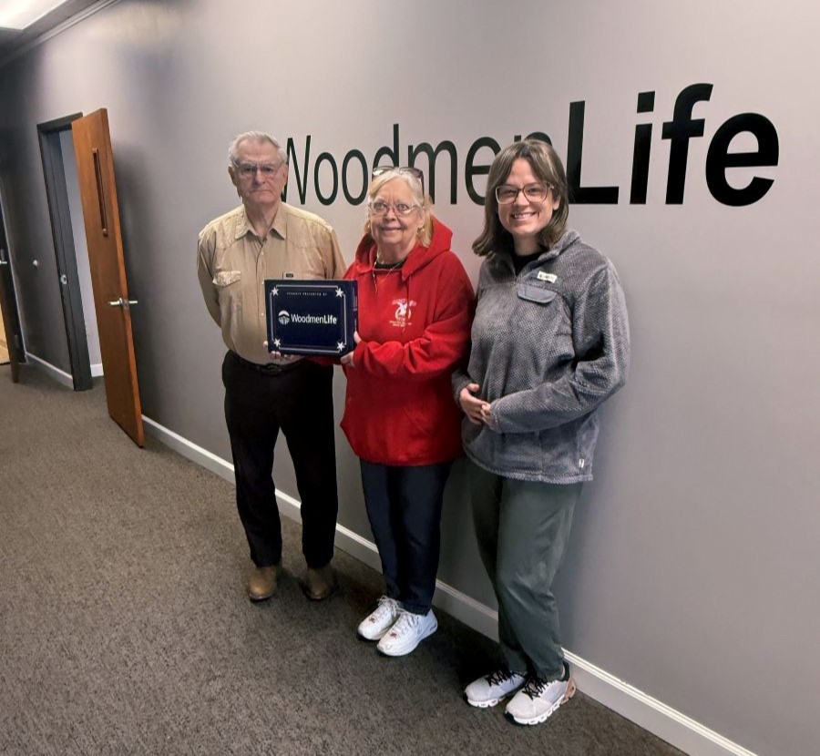 Three people stand together. The woman in the middle is holding a navy blue box that has the WoodmenLife logo on it. The WoodmenLife name is also on the wall behind the people.