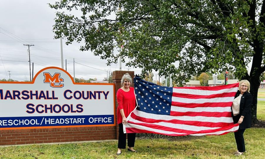 Two women stand outside holding an American flag. They are standing next to a sign that says "Marshall County Schools Preschool/Headstart Office."