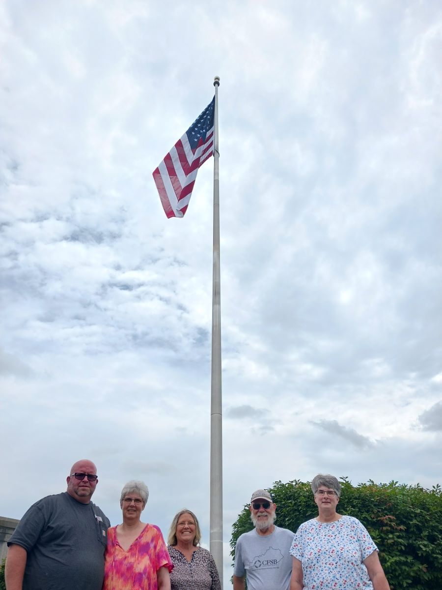 Five people stand in front of a flag pole, on top of which is waving the newly donated flag.