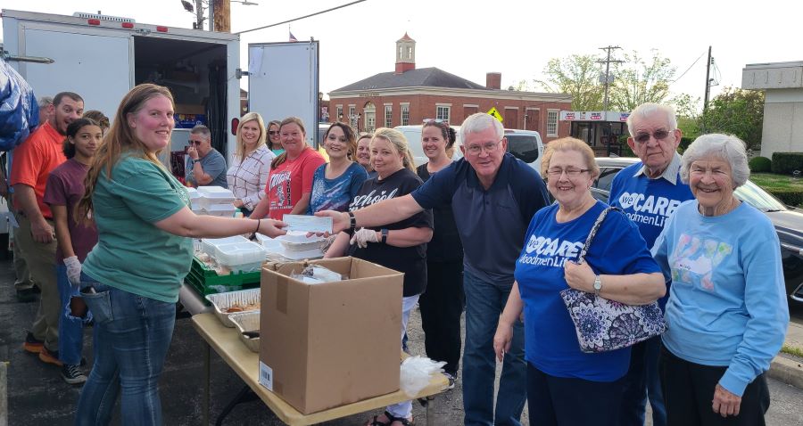 A group of people -- about 17 total -- are standing outside around a table. On the table are white takeout boxes. In the foreground, two people are exchanging a check.