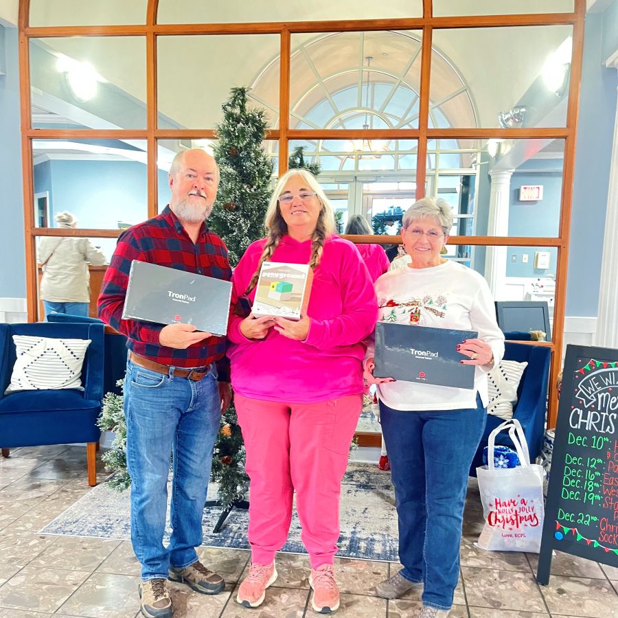 Three people stand together in the photo. They are each holding a box. Behind them is a Christmas tree.