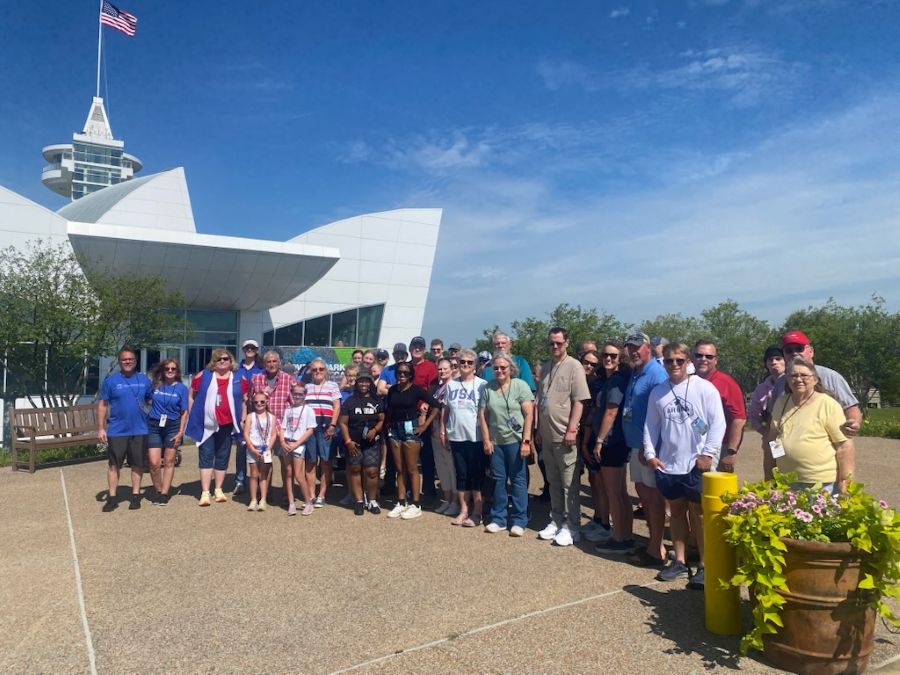 Photo shows a group of about 35 people. They are standing outside of a building. There is a large potted plant next to the group, and there are trees and blue sky behind them.