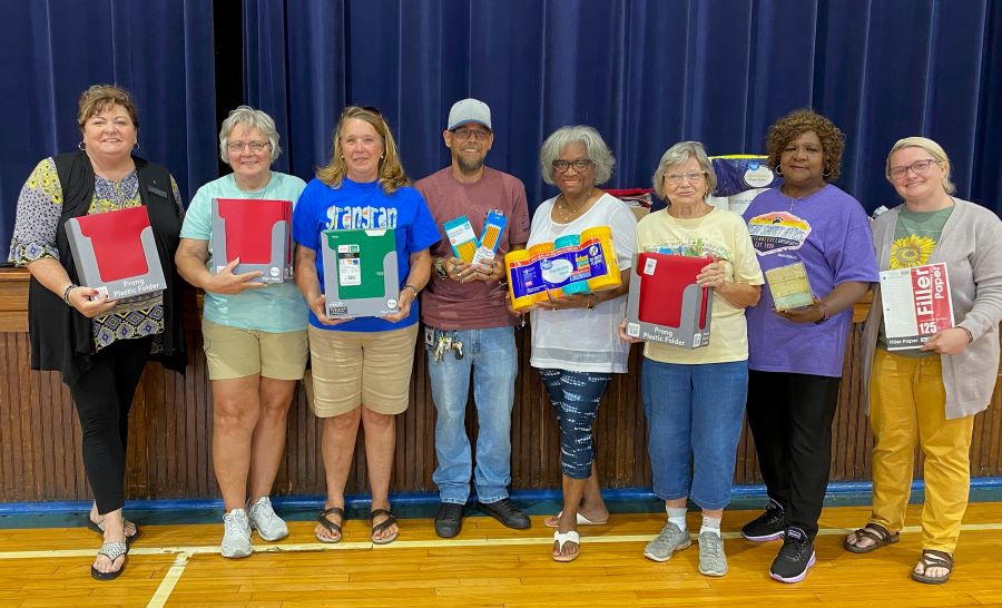 Eight people stand together for a photo. They are holding folders, pencils, Lysol wipes and other chapter donations for the elementary school.