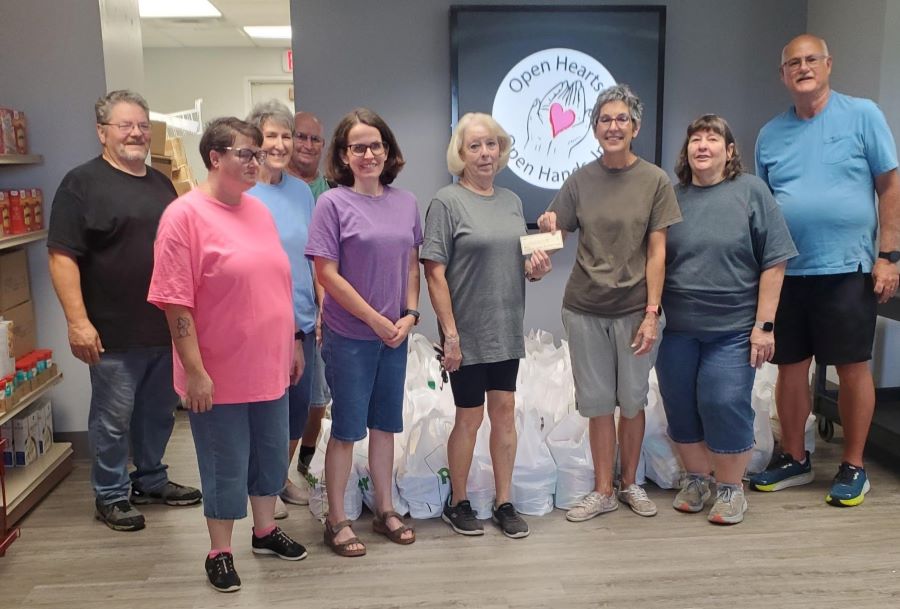 Nine people stand together for a check presentation. Behind them on the wall is the Open Hearts Open Hands, Inc. logo. On shelves along the wall and in bags near the group are donated items.