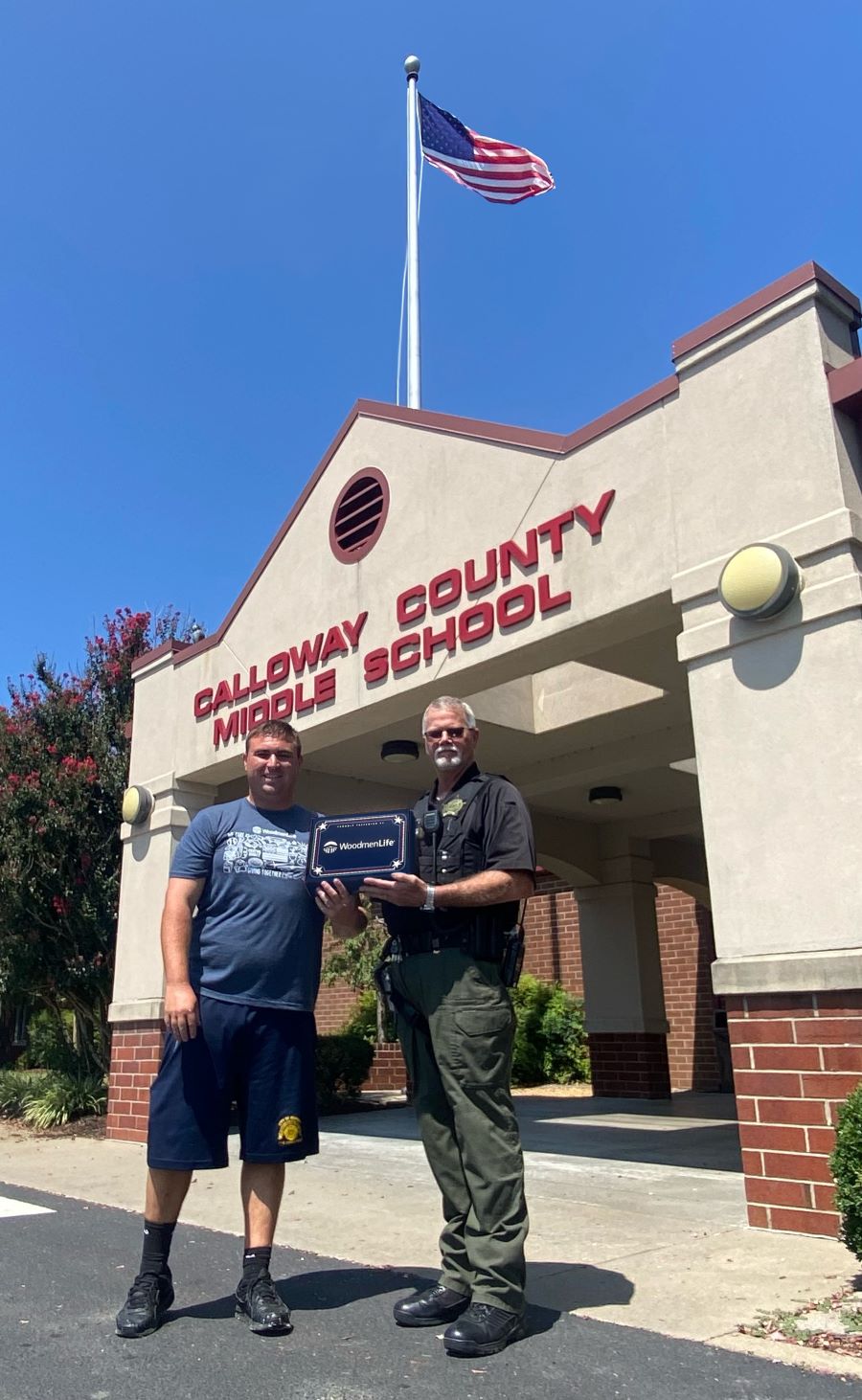 Two men stand together holding a flag box. Behind them is a school, Calloway County Middle School. The photo is taken from the ground level looking up, so you can see the flag atop the school.