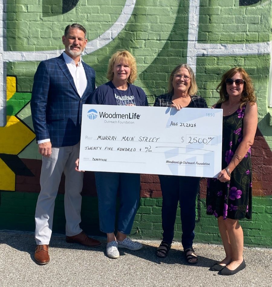 Four people stand together holding an oversized check. The check is made out to Murray Main Street in the amount of $2,500.