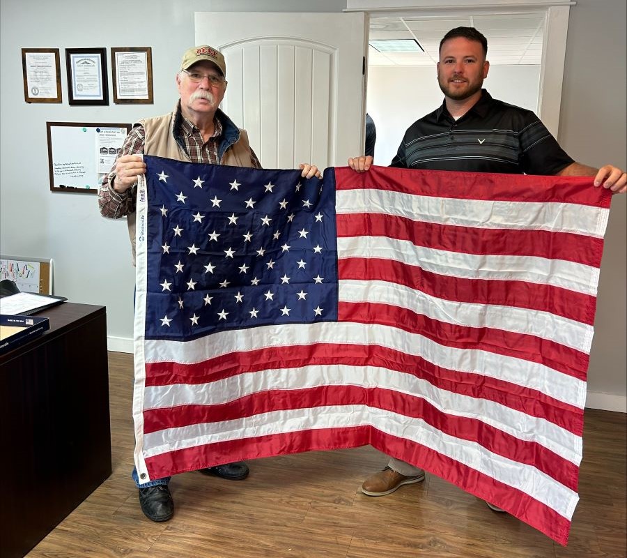 Two men are standing in an office holding an American flag.