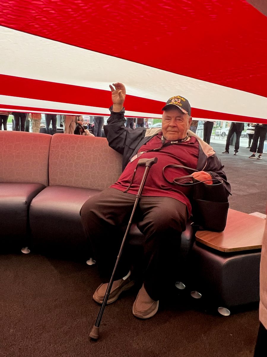 A man is seated, and an oversized American flag is being displayed over his head. He is helping hold up the flag with his right hand, and holding a bag with his left hand. There is a cane resting in front of him, and he is wearing a "Disabled Veteran" baseball hat.