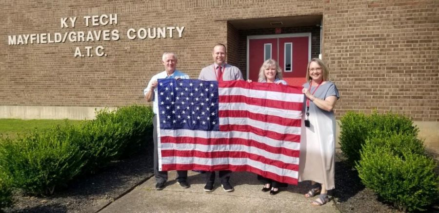 Four people stand together holding an American flag. Words on the building behind them say "KY Tech, Mayfield/Graves County A.T.C."