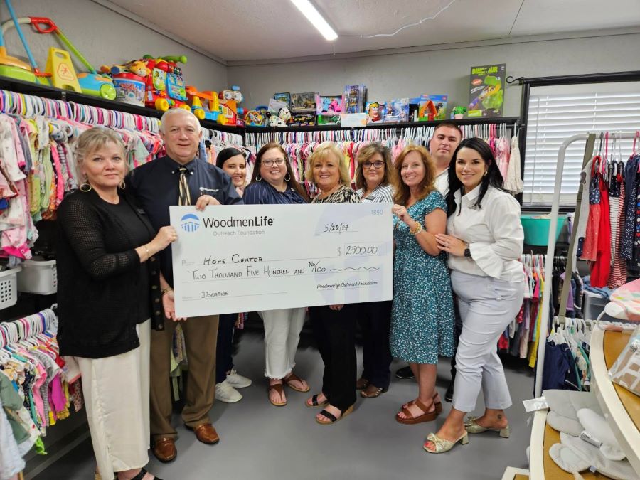 A group of nine people stand together holding an oversized check. The check is made out to the Hope Center and worth $2,500. Behind them are brightly colored children's toys and clothing.