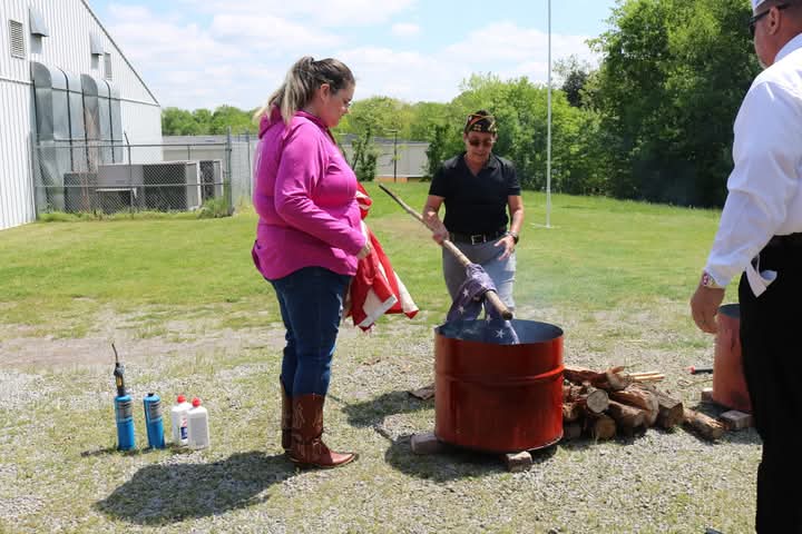 Three people stand around a metal fire pit. On the ground near them is chopped wood and lighter fluid. They are using a long stick to lower an American flag into the fire pit as part of a ceremonial retirement.