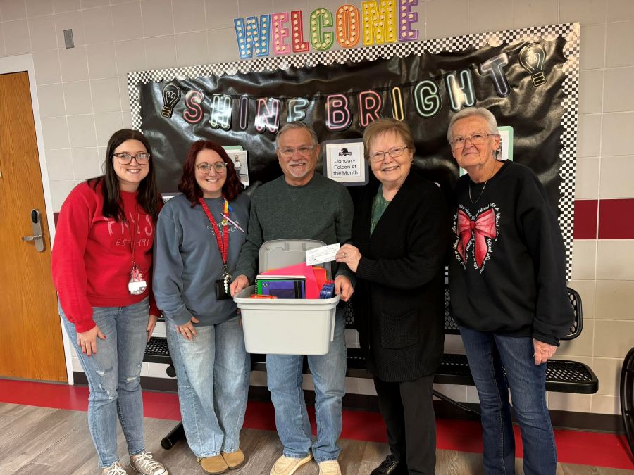 Five people stand together for the photo. The man in the middle is holding a box with notebooks and other school supplies. The woman to the right of him is holding a check. They are standing in a school hallway, and behind them is a bulletin board that says "Welcome" and "Shine Bright."