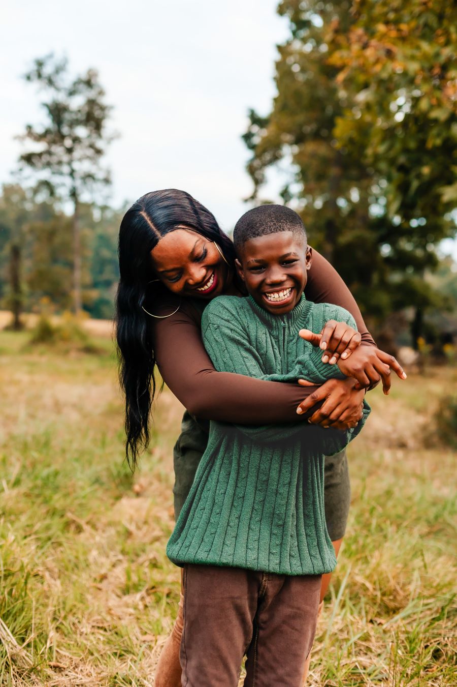 WoodmenLife member Iesha Kelley and her 12-year-old son pose for a photo. They are outdoors, and there is native grass and trees behind them. She his standing behind him, hugging him.