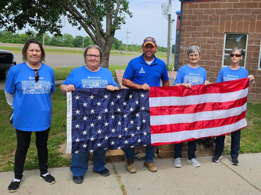 Five people are standing outside and holding an American flag. All five people are wearing blue shirts.