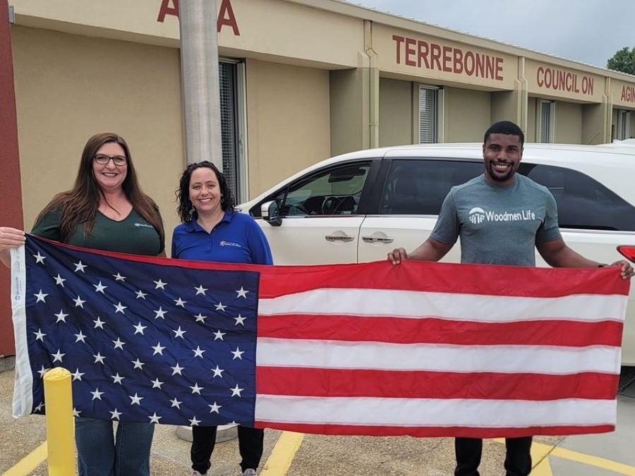 Three people hold an American flag. They are standing outside a building that says Terrebonne Council on Aging.