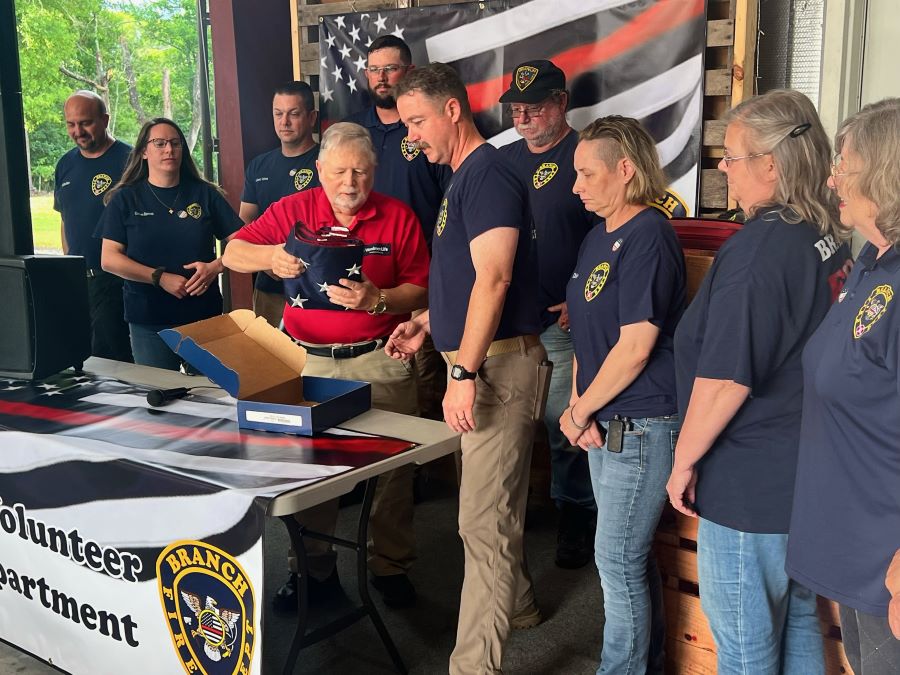 A WoodmenLife representative, wearing a red polo shirt with the WoodmenLife logo, presents a folded-up American flag to a group with the Branch Volunteer Fire Department. They are all wearing navy blue T-shirts with the Fire Department logo on them. The open box that the flag came out of is on a table in front of the group.