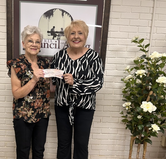 Photo shows two women standing together holding a check. Behind them is a white brick wall, and beside them is a plant with white flowers. On the wall behind them is a framed image that says "Iberia Council on Aging."