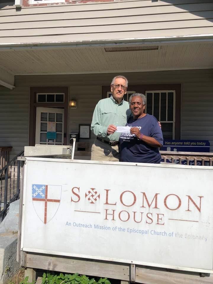 A man and a woman stand together holding a check. In front of them is a sign that says "Solomon House" and behind them is the front door and front porch of a building.
