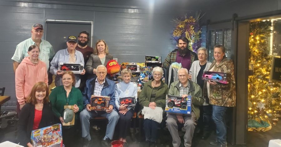 Fourteen people are in the picture — some seated and some standing. They are posing with toys they are donating. There is a Christmas tree behind them.