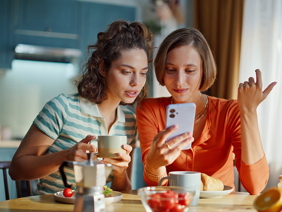 Stock photo shows two young women. They are sitting at a table together and looking at a cellphone. One woman is holding a coffee cup, and there are plates with food on the table in front of them.