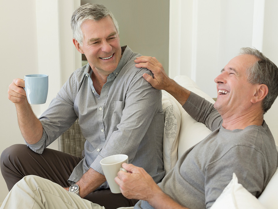 Stock photo shows two men sitting on a couch. They are smiling and laughing, and one man has a hand on the other man's shoulder. They are both holding coffee cups.