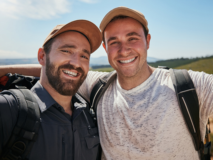 Stock photo shows two men with arms around each other, looking at the camera and smiling. They are both wearing hats and backpacks. They are outside, with blue sky and landscape behind them.