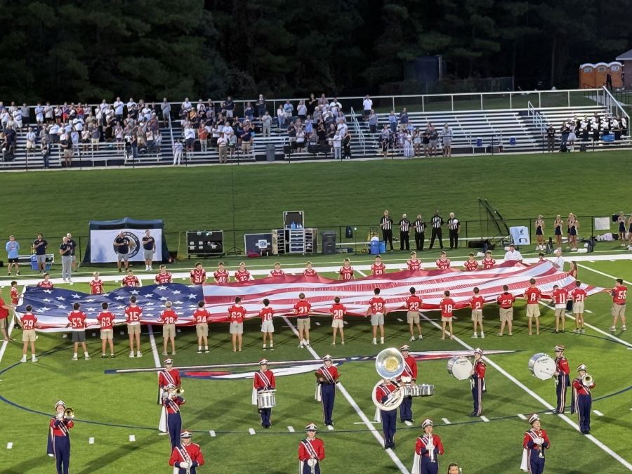 Photo shows an oversized American flag being held on display on a football field. Students in red football jerseys are holding the flag as a marching band stands in front of them. Spectators in the bleachers and on the sidelines watch.