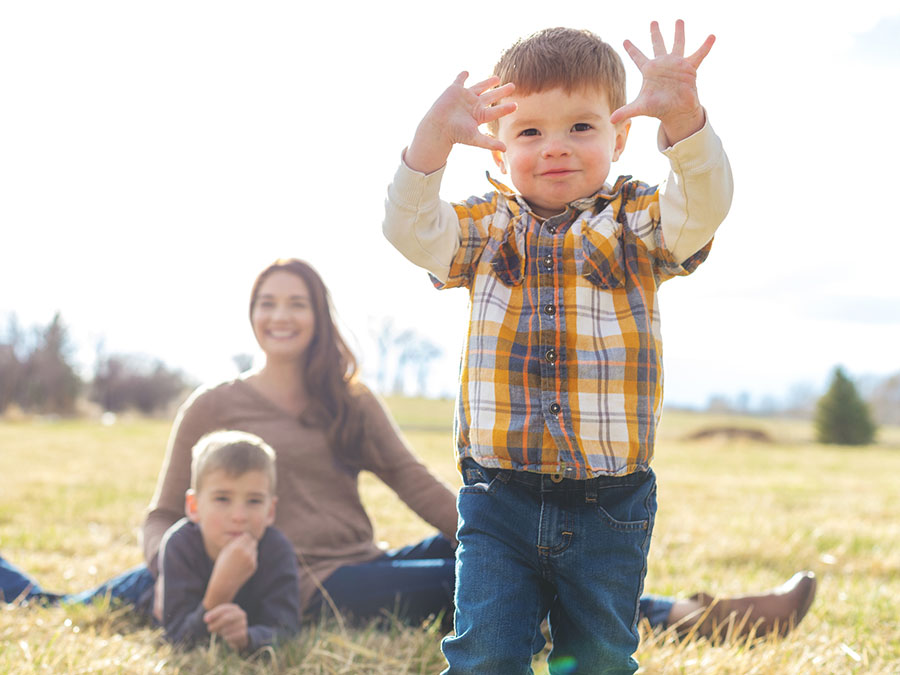 Stock photo shows a woman and two children. One child is in the foreground, walking toward the camera with his hands up. The woman and the other child are seated in the background, looking on. They are outside in a grassy field.