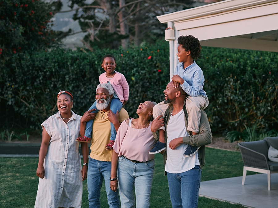 Stock photo shows six people. Two men and two women are standing together, and there are two children, each one sitting on a man's shoulders. Everyone is smiling.