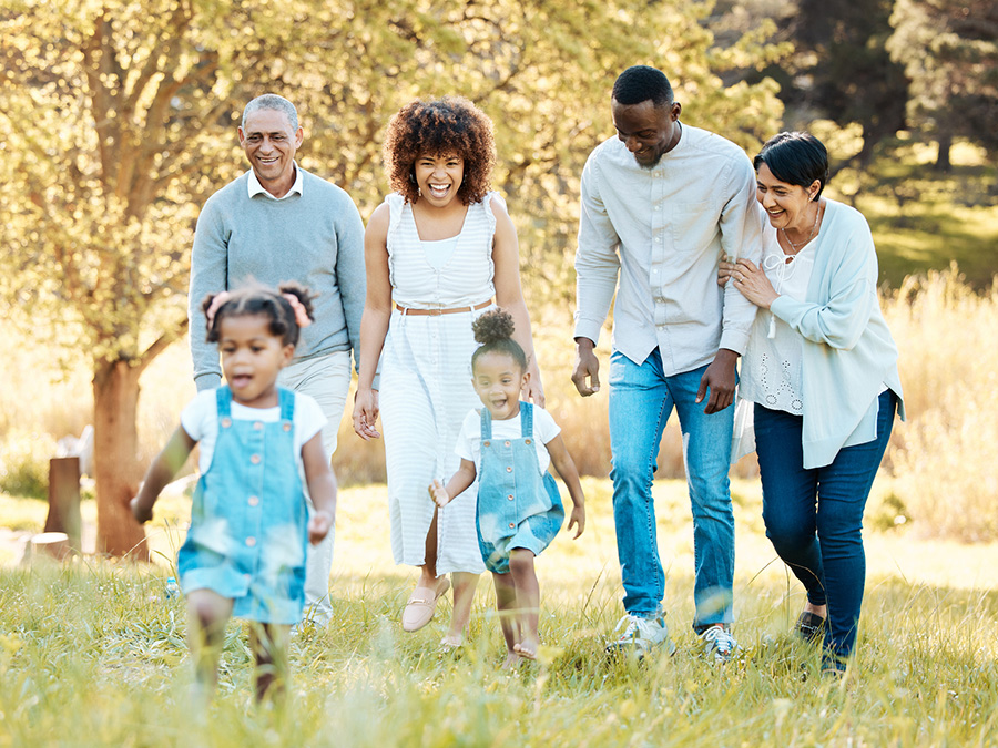 Stock photo shows six people. Two young girls are in the foreground running through knee-high grass. Behind them are four adults: a middle-aged man and woman, and an older man and woman. They are smiling as they watch the children in front of them.
