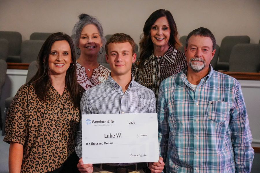 Photo shows five people standing together, posing for the photo. At the center is a young man holding an oversized WoodmenLife-branded check. The check is made out to Luke W. in the amount of $10,000. Flanking the young man are a man and woman, his parents. The last two people -- two women standing behind the family -- are WoodmenLife associates who presented the scholarship check.