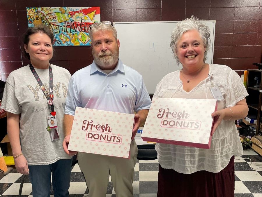 Three people pose for the photo. The person in the center and on the right are holding up boxes that say "Fresh Donuts."