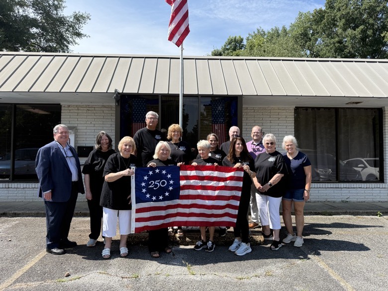 A group of 13 people stand together in the photo. They are holding a commemorative America 250 flag, which has 13 stars in a circle around "250" in the upper-left blue square. The group is standing in a parking lot, and there is a building behind them.