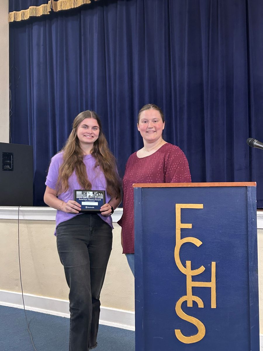 A woman stands next to a podium with a high school-aged girl. The girl is holding an American History Award plaque. Next to them is a podium that says "FCHS," and behind them is a stage with dark blue curtains.