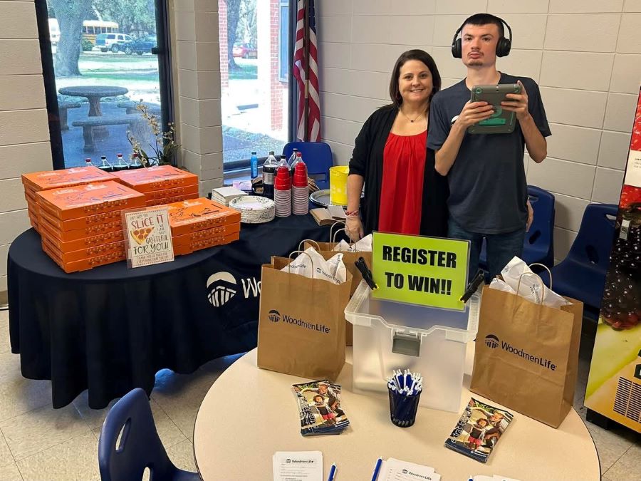 A woman and a young man pose for the picture. Behind them is a table holding at least 20 boxes of pizza, as well as a stack of plates, cups, and 2-liter bottles of pop. On the round table in front of them are WoodmenLife-branded bags, WoodmenLife flyers, and a box where people can enter a drawing for prizes.