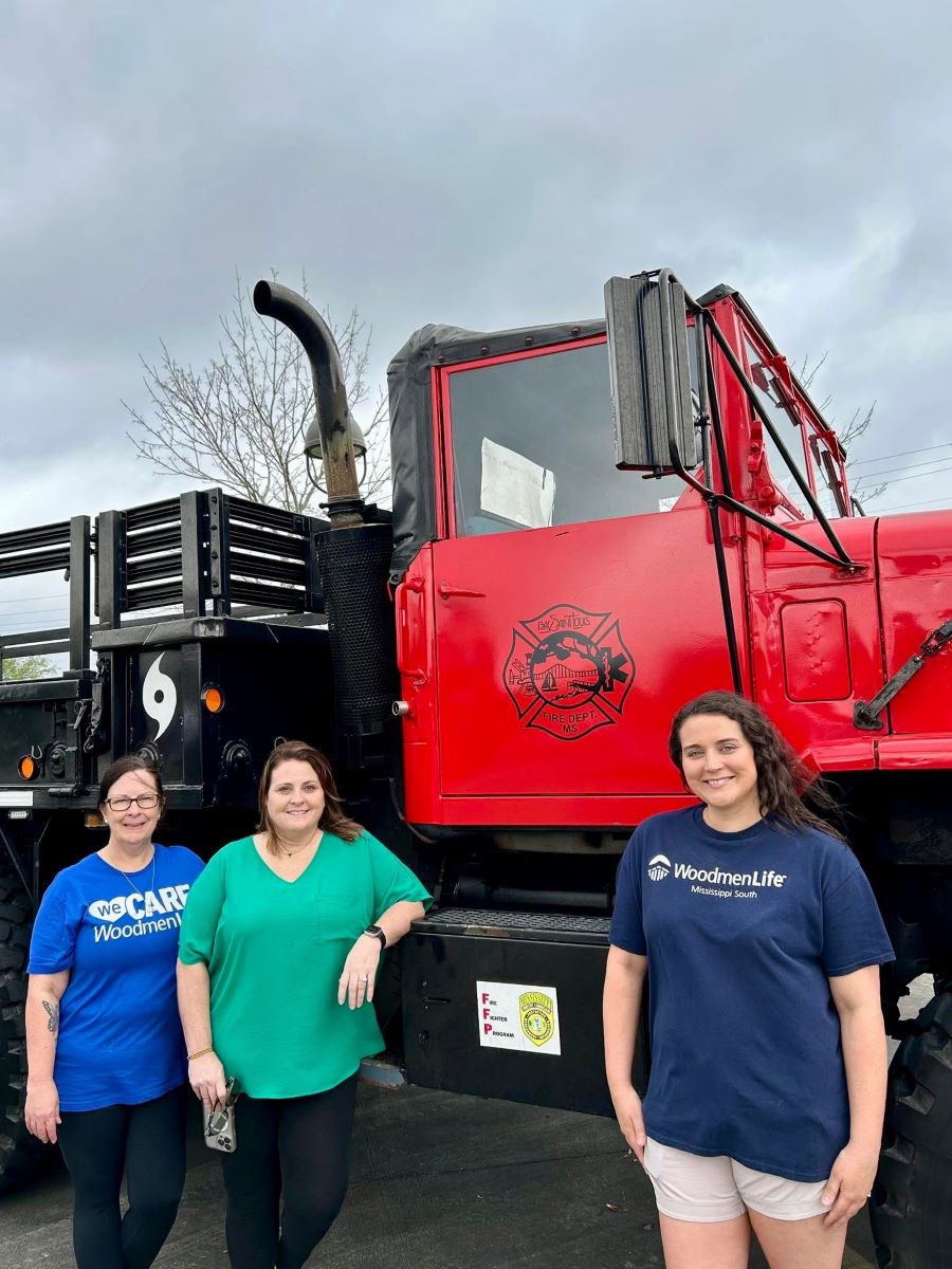 Three women smile for the photo in front of a large red truck. The woman on the left and right are wearing WoodmenLife-branded T-shirts. Behind them, the sky is dark and cloudy.