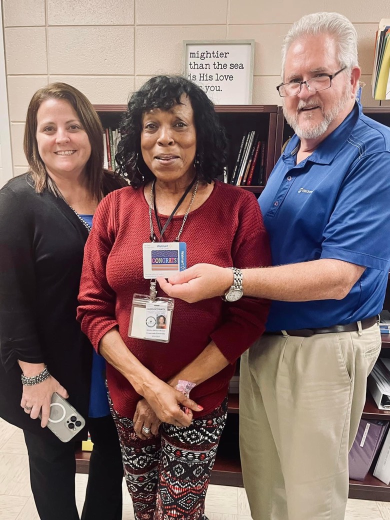 Three people are in the photo. The woman in the center is a teacher who is being presented with a Walmart gift card.