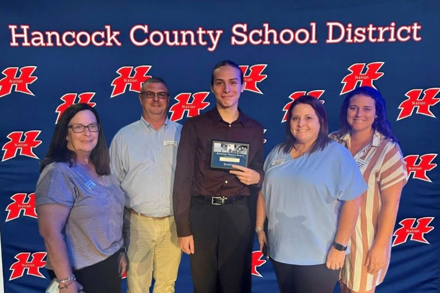 Five people appear in the photo. In the middle is a high school student holding a plaque that says "American History Award." The backdrop says "Hancock County School District" and has the district's "H Nation" logo.