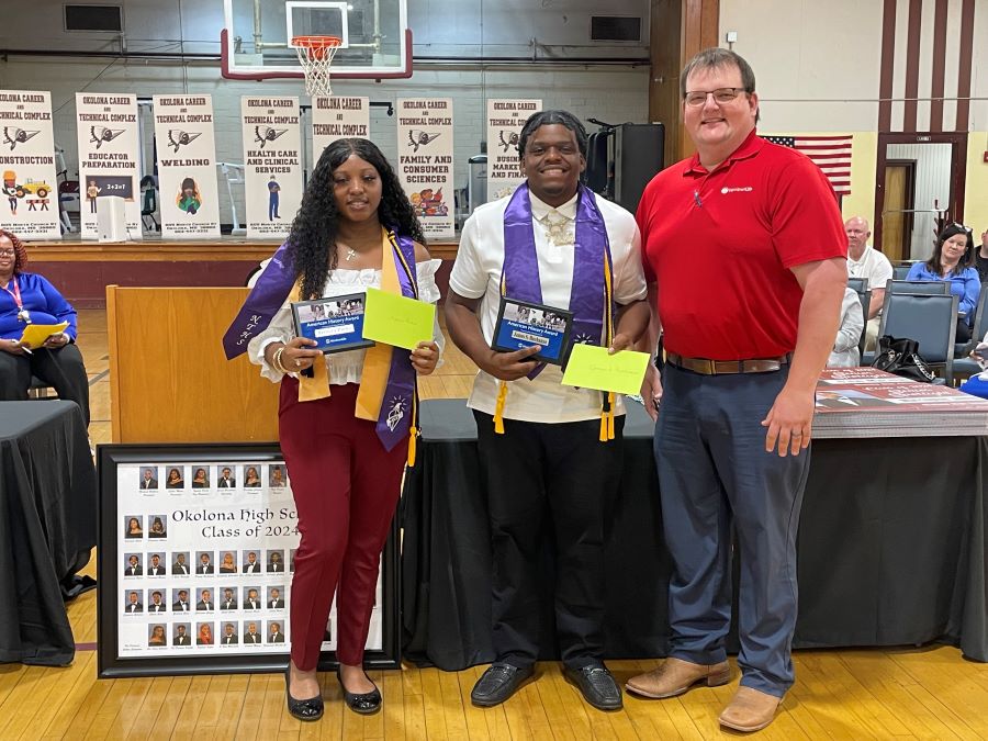 Three people stand together for the photo. The person on the left and in the center are graduating high school students. They are each holding plaques commemorating their American History Awards from WoodmenLife. The person on the right is wearing a red polo shirt with the WoodmenLife logo. The group is in a school gym.