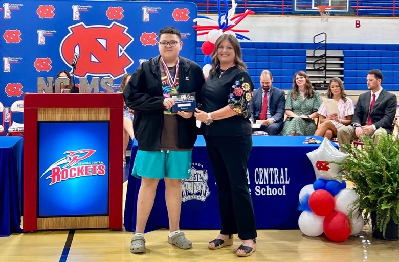 A woman presents a plaque to a middle school student. Behind them is a blue-and-red Neshoba Central Middle School-branded backdrop, podium, and table with table cloth.