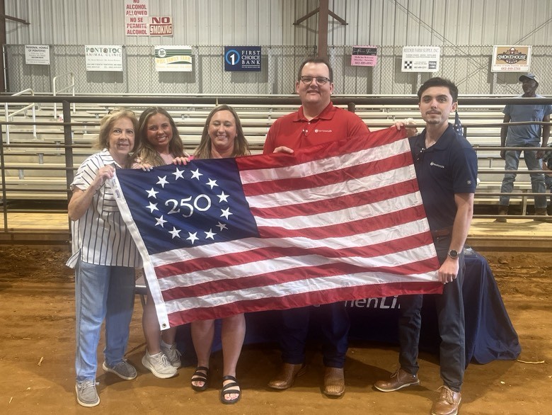 Five people stand together holding a commemorative America 250 flag, which features a circle of 13 stars around "250" in the upper-left blue square. Rows of bleachers are behind the group.