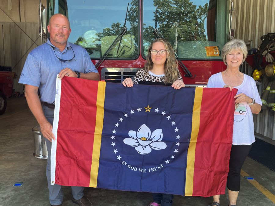 A man and two women stand in front of a fire truck holding a Mississippi state flag.