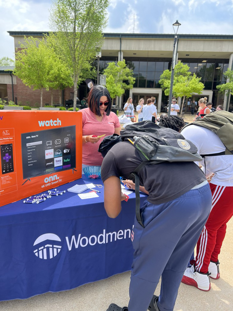 A table with a WoodmenLife-branded tablecloth is set up outside on a college campus. On the table is a television box and other giveaways. A woman is standing behind the table, and two students wearing backpacks are bending over to fill out a form at the table.
