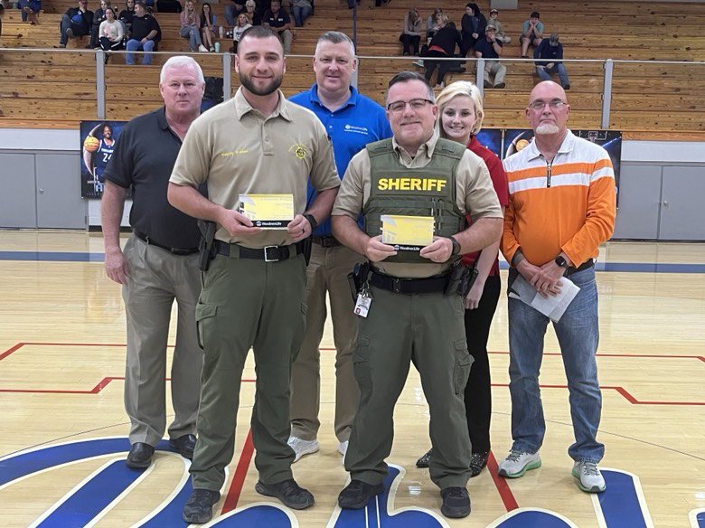 Six people are standing in a school gym. The two people closer to the front are wearing law enforcement uniforms and are holding their awards.