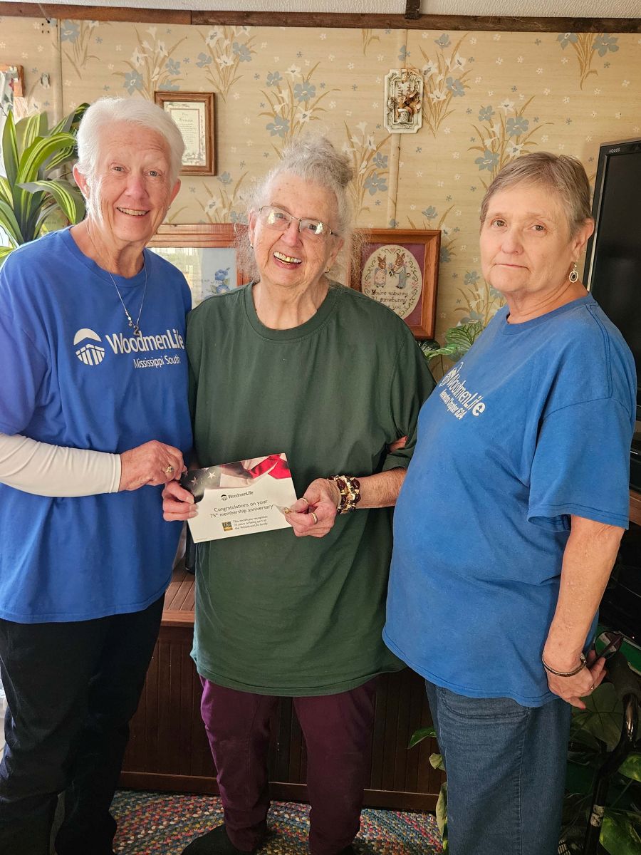 Three women appear in the photo. The two women on either end are wearing blue WoodmenLife T-shirts. The woman in the middle is holding a certificate.
