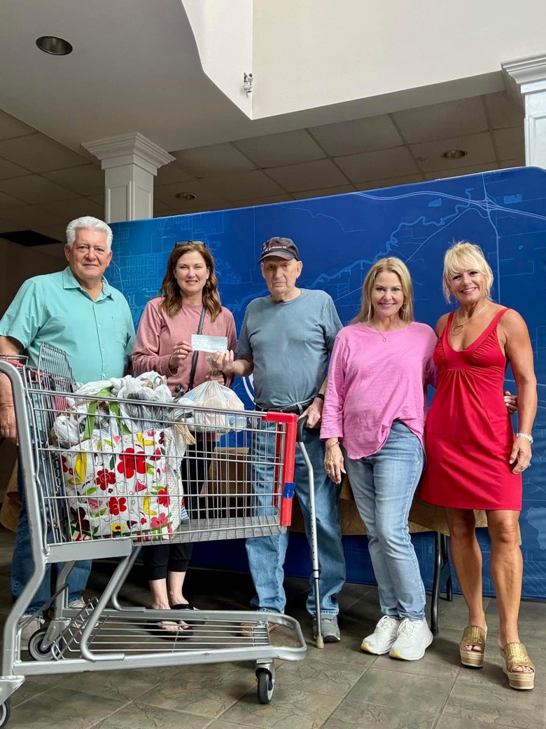 Five people stand together. Two people in the middle are exchanging a check, which is a $500 donation to the Backpack Buddies program. In front of the people is a grocery cart with bags inside.