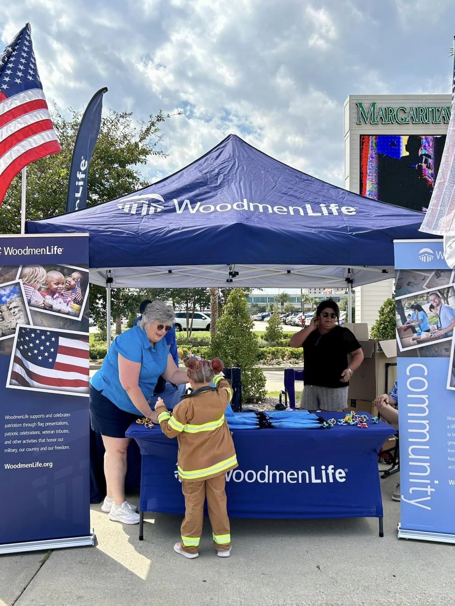 A WoodmenLife-branded tent and table are set up at an outdoor event. A young girl wearing a firefighter's uniform is at the table, talking with the WoodmenLife volunteers.