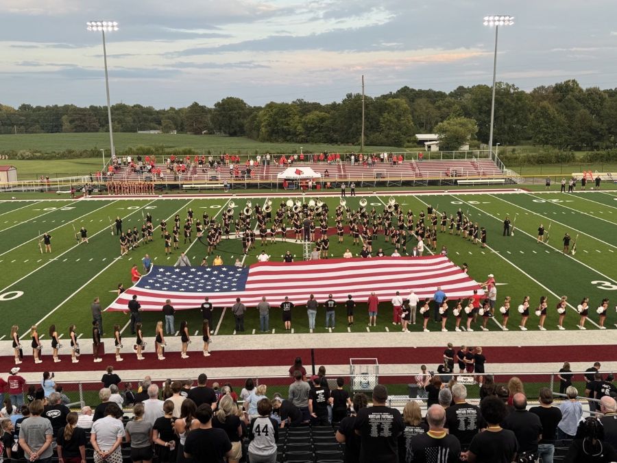 Photo shows an oversized American flag displayed on a football field. A marching band is on the field beyond the flag. Spectators are watching in the near bleachers and from the opposite side of the field.